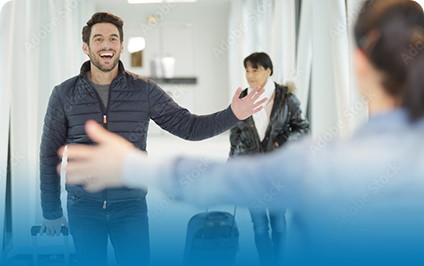 Man smiling and extending arms for a warm welcome at an airport arrival gate with luggage and another traveler