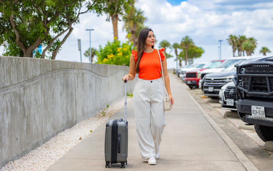 A young woman in a bright orange shirt walks in the airport parking lot with her luggage.