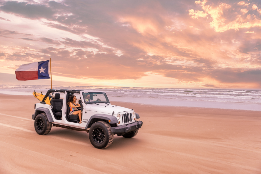 The beach during sunset, with two people driving a white Jeep with a Texas Flag waving in the back.